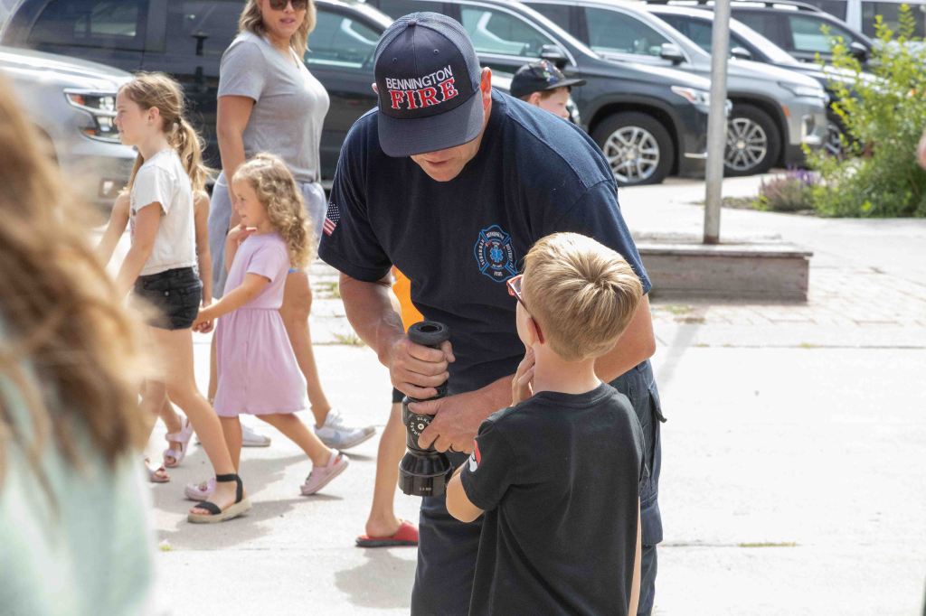 A firefighter interacting with a young boy, demonstrating a fire hose, while a group of children and adults are present in the background.