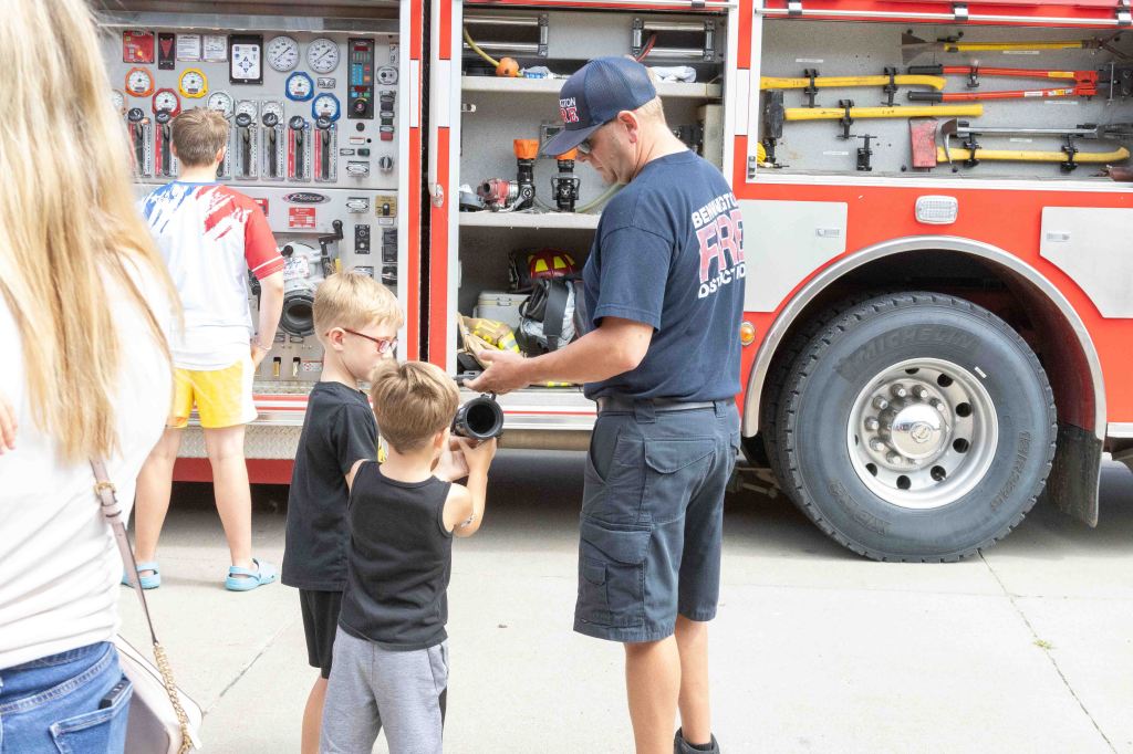 A firefighter demonstrates equipment to two young boys next to a fire truck, while another child observes in the background.