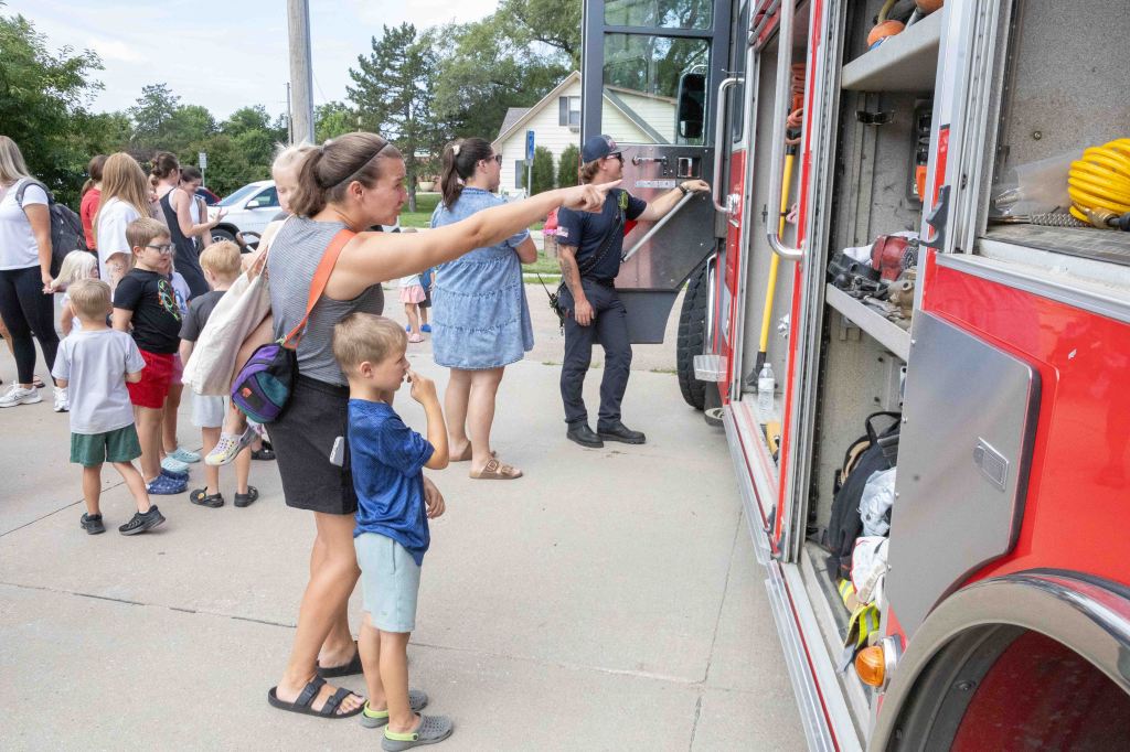 A group of children and adults line up by a fire truck, engaging with a firefighter who points out equipment. A woman and a child stand in the foreground, looking curiously at the truck's contents.