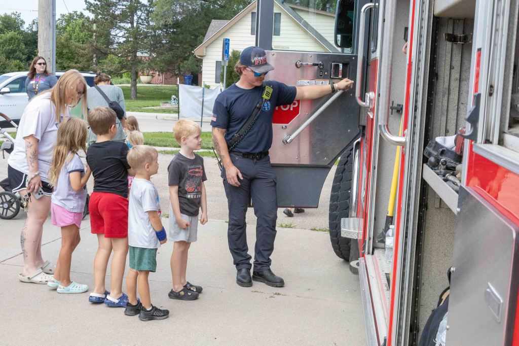 A firefighter interacts with a group of children in front of a fire truck, with parents observing in the background.