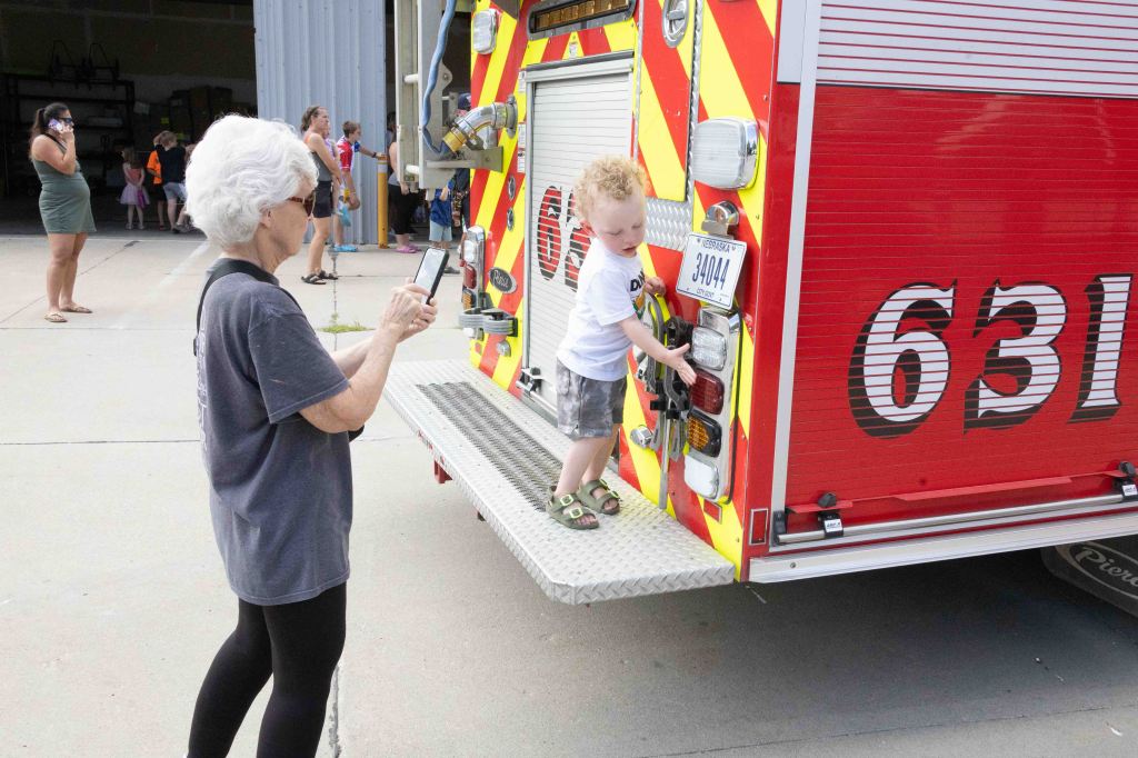 An older woman with gray hair taking a photo of a young child climbing on a fire truck with colorful paint, while other people gather in the background.