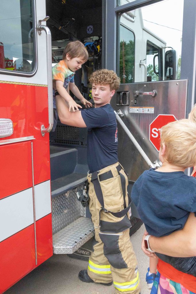 A firefighter helping a young child step down from a fire truck, with another child watching from the side.