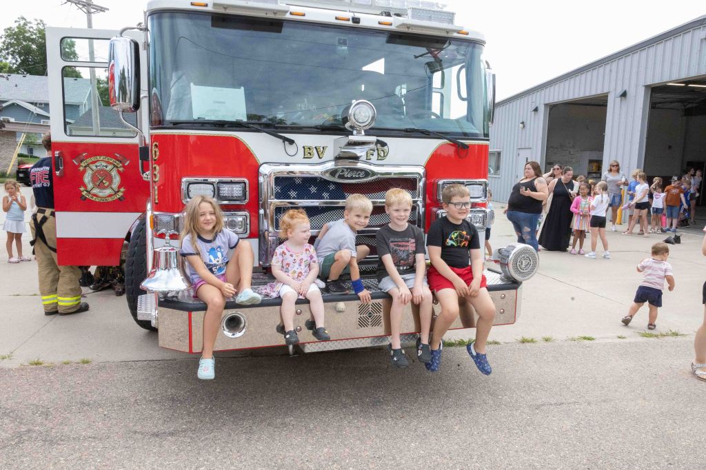 A group of five children sitting on the front of a fire truck, smiling and enjoying an event with a crowd in the background.