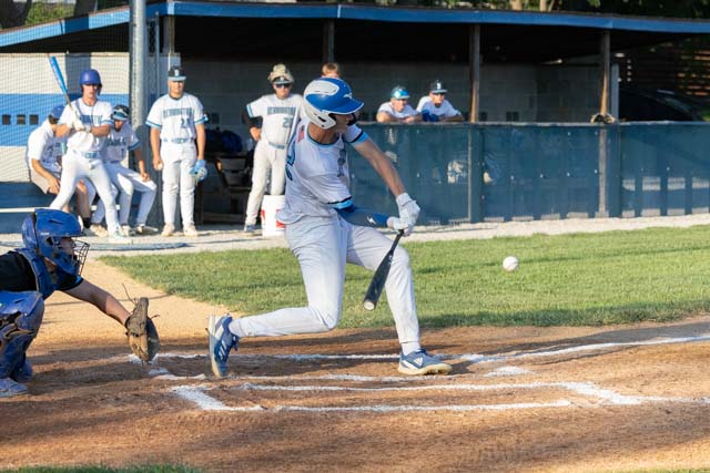 A baseball player swings at a pitch while a catcher crouches in the foreground, with teammates visible in the background.