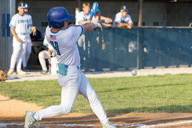 A young baseball player in a blue helmet and white uniform swings at a pitch during a game, with teammates visible in the background.