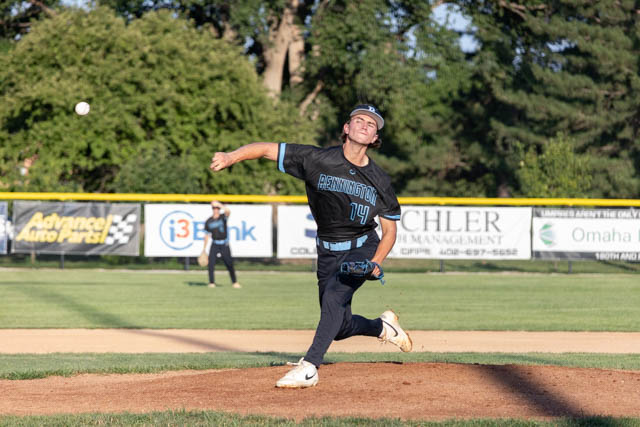 A young baseball pitcher in a blue and black uniform throws a pitch from the mound during a game.