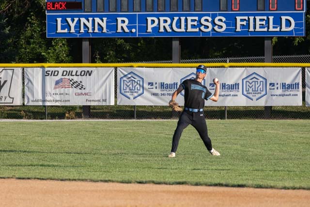 A baseball player in a black and blue uniform stands in the outfield at Lynn R. Pruess Field, preparing to catch a ball, with a scoreboard displaying 0-0 in the background.