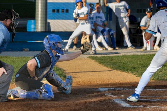 A baseball catcher in protective gear is crouching ready to receive a pitch, while a batter prepares to swing. In the background, team members are watching from the dugout.