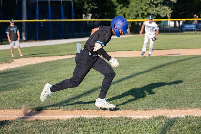 A baseball player in a blue helmet and black uniform running toward first base on a grassy field, with another player preparing to field the ball in the background.