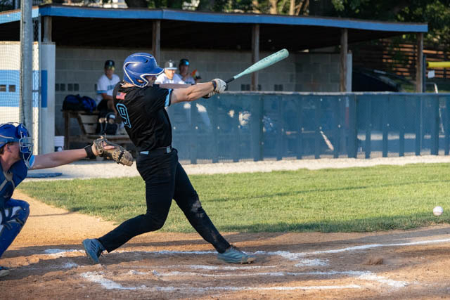A baseball player swings a bat at home plate during a game, with another player in a catcher's gear behind him.