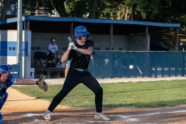 A baseball player in a black uniform and blue helmet is preparing to hit a ball during a game, with a catcher in blue gear behind him and a dugout visible in the background.