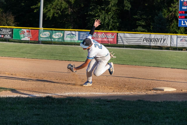 A baseball player in a white uniform diving to catch a ball on the infield, with the sun setting in the background.