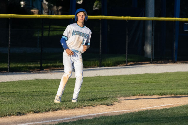 A baseball player in a white and blue uniform, wearing a helmet, jogging along the base path on a diamond with green grass and a yellow outfield fence in the background.