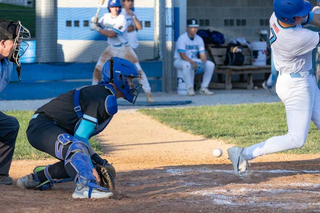A baseball player swings at a pitch while a catcher prepares to receive the ball. In the background, teammates watch from the dugout.