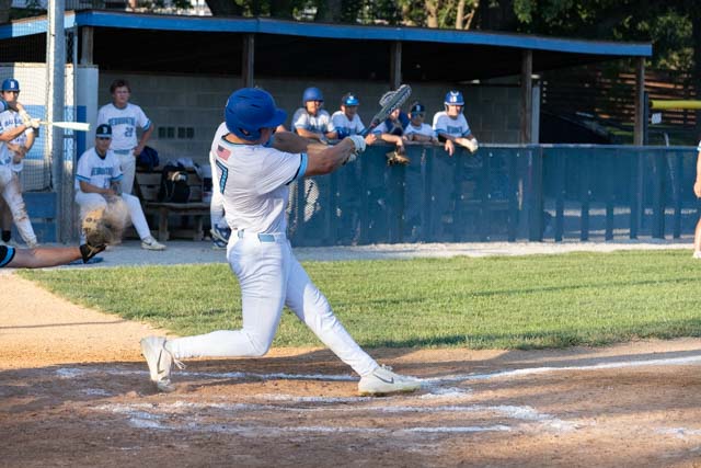 A baseball player in a white uniform swings a bat on the field while teammates observe from the dugout in the background.