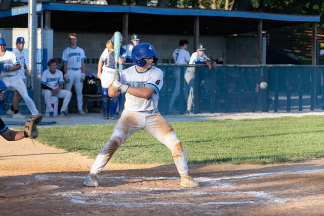 A baseball player in a white and blue uniform stands ready to bat, with a baseball approaching from the left side of the image. The player is focused and in a batting stance. In the background, other players are visible sitting on a bench.