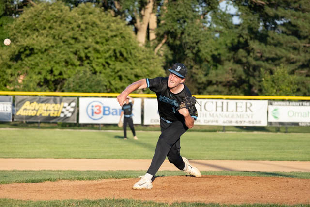 A baseball pitcher throwing a ball on a field, wearing a black jersey and cap, with a focused expression.