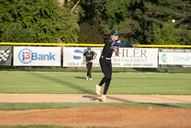A baseball player in a black uniform throws the ball while standing on the infield, with another player positioned in the background. The scene is set on a baseball field with advertisements visible along the outfield fence.