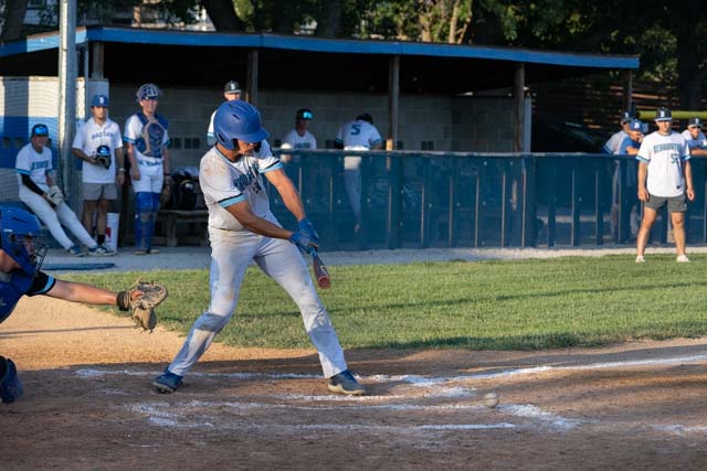 A baseball player swinging a bat to hit a pitch on a sandy field, with teammates in the background watching from the dugout.
