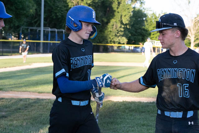 Two young baseball players in black uniforms with blue accents celebrate a play with a fist bump on the field.