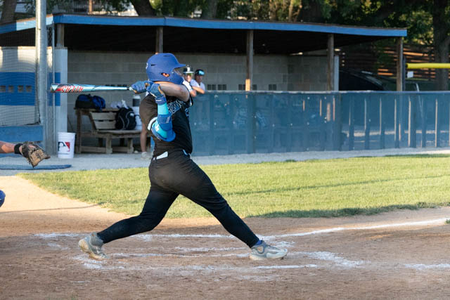 A young baseball player swinging a bat at home plate during a game, with a grassy field and dugout in the background.