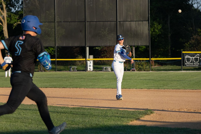 A baseball player in a white uniform throws the ball towards a teammate while another player in a black uniform runs towards first base.