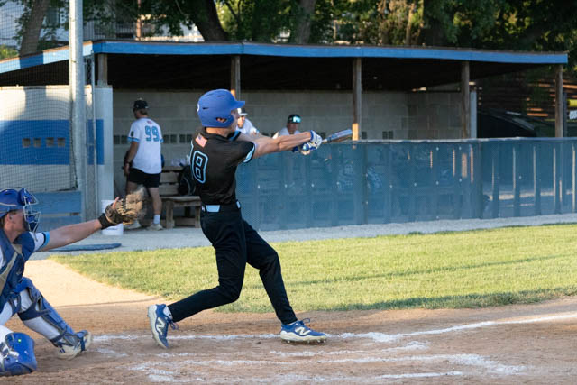 A young baseball player in a black uniform swings at a baseball during a game, with a catcher in blue gear ready to catch the pitch.