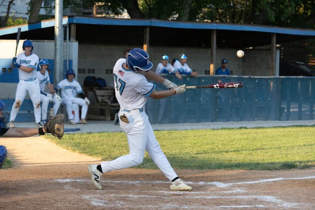 A baseball player swings at a pitch during a game, captured in mid-action. Teammates are seated in the dugout in the background.