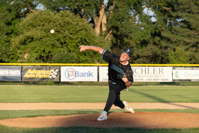A baseball player in a black uniform is pitching on a sunny field, with trees and advertisements in the background.