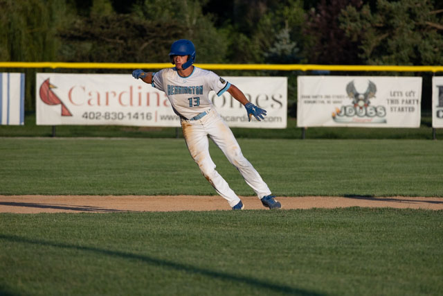 A young baseball player in a blue helmet and jersey runs towards first base on a baseball field, with advertisements and greenery in the background.