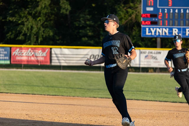 A young baseball player in a black uniform with the number 12, celebrating on the field after a play. The background shows a scoreboard with game details.