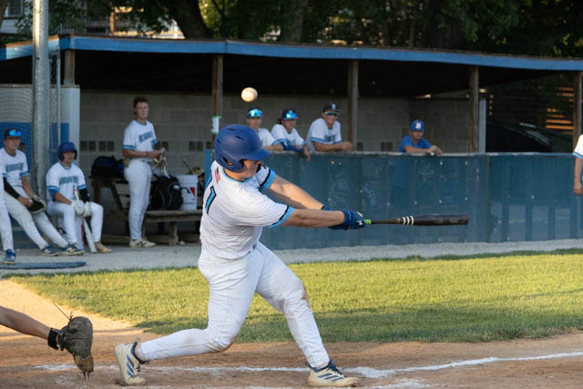 A baseball player in a white uniform is swinging a bat to hit a pitched ball during a game, with teammates in the dugout in the background.
