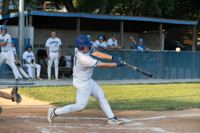 A baseball player in a light blue uniform swings a bat at a pitch during a game, with teammates sitting in the dugout in the background.