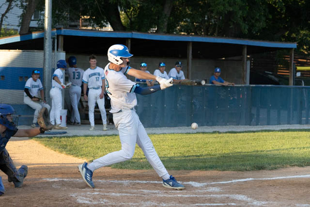 A baseball player in a white uniform swings a bat during a game, with teammates in the dugout watching in the background.