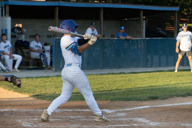 A baseball player in a blue helmet and white uniform is swinging a bat in a game setting, with teammates and coaches visible in the background.