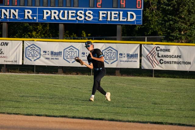 A baseball player wearing a black jersey and cap fields a ball on a grassy field in front of a scoreboard at Prueess Field.