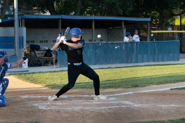A baseball player in a black uniform prepares to swing a bat at a pitched ball during a game, with a catcher in blue gear behind him.