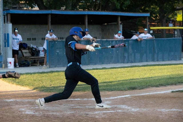 A baseball player in black uniform swings a bat during a game, with teammates in the background watching from the dugout.