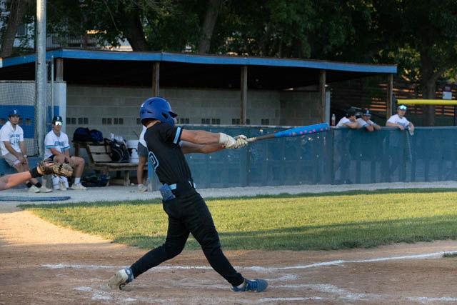 A baseball player swinging a bat during a game, with teammates in the background watching from the dugout.