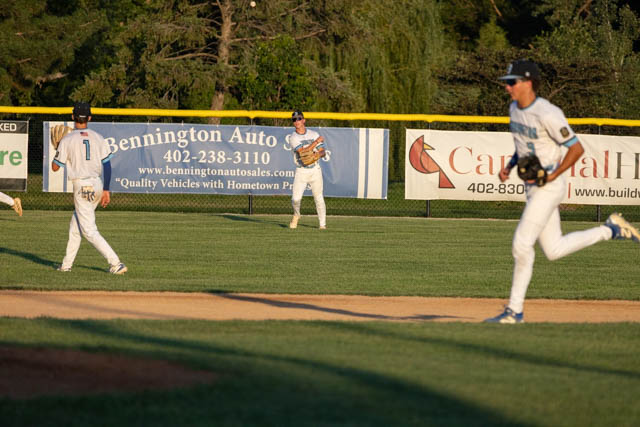 Three young baseball players on the field during a game, with the outfield fence and advertisements in the background.