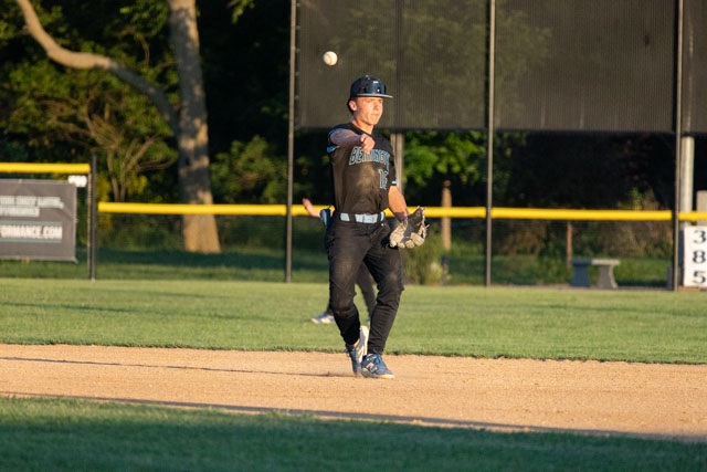 A young male baseball player in a black uniform throws a baseball while standing on the infield of a baseball diamond during a sunny evening.