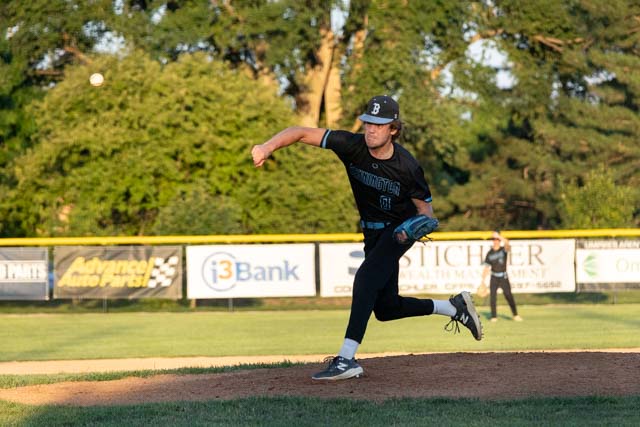 A baseball pitcher throwing a pitch on a field during a game, with trees and a scoreboard in the background.