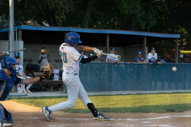 A youth baseball player swings a bat at a pitched ball during a game, with players in the dugout visible in the background.