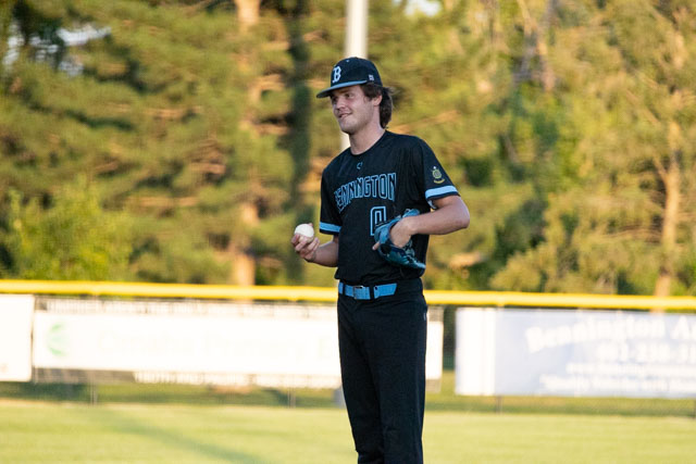 A young baseball player stands on the field, holding a baseball and a glove, wearing a black and blue uniform with the name 'Bennington' on it.