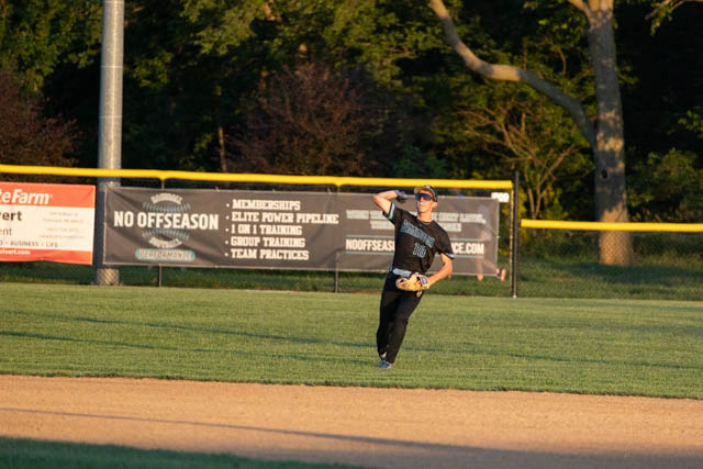A baseball player in a black uniform catches a fly ball during a game, with a yellow outfield fence and a banner in the background.