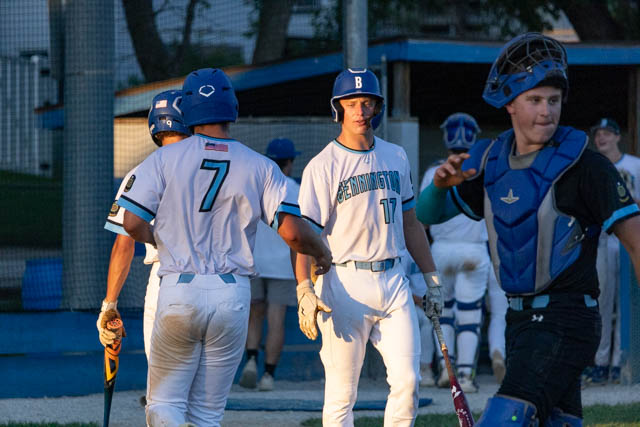 Two baseball players in white uniforms congratulate each other on the field, with one player wearing the number 7 and another wearing number 17. A catcher in blue gear is visible in the foreground.