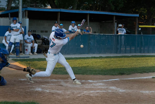 A baseball player wearing a blue helmet and a white jersey with the number 7 swings at a ball during a game, while a catcher crouches behind home plate.