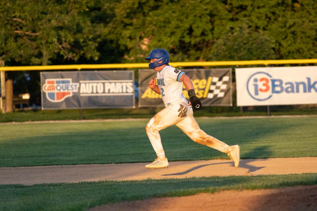 A baseball player in a white uniform with blue accents runs on the field during a game, showing signs of dirt on his pants as he heads towards the base.
