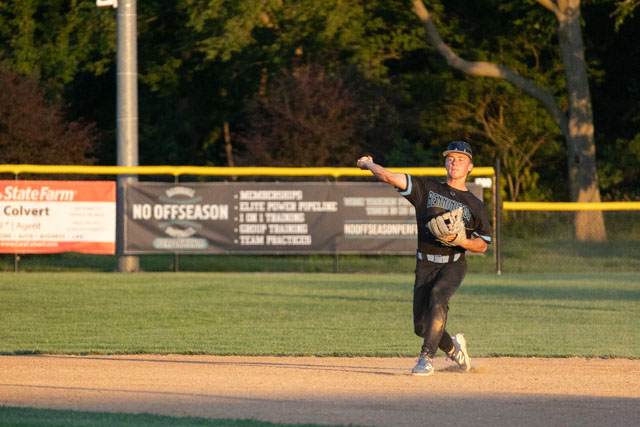 A baseball player in a black uniform throwing a ball while standing on the infield during a sunset.