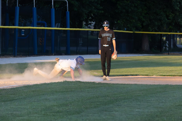A baseball player dives into a base while a second player stands nearby, watching. Dust rises from the field.